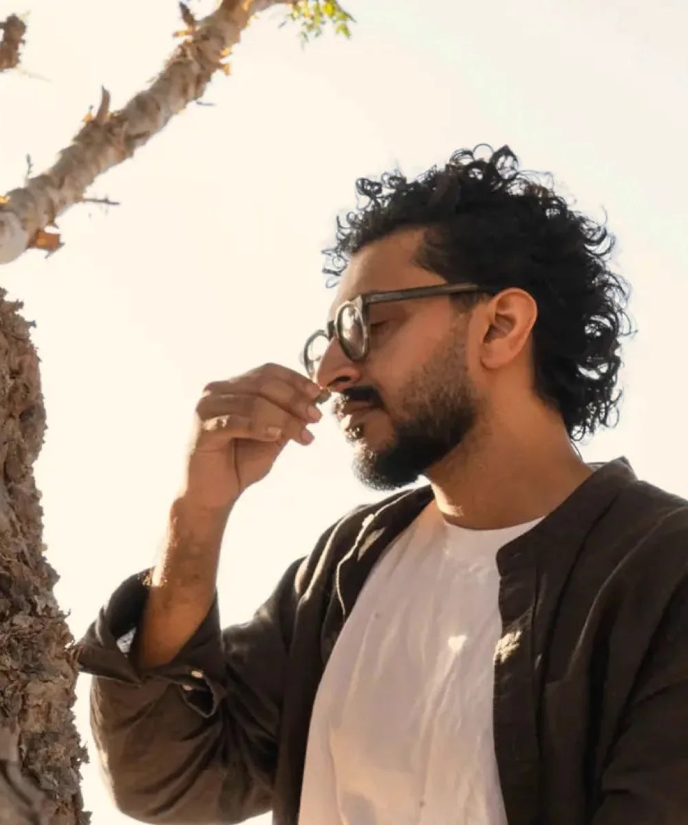 Sho, eyes closed, holds a piece of frankincense resin to his nose beside the trunk of a Boswellia tree in Dhofar